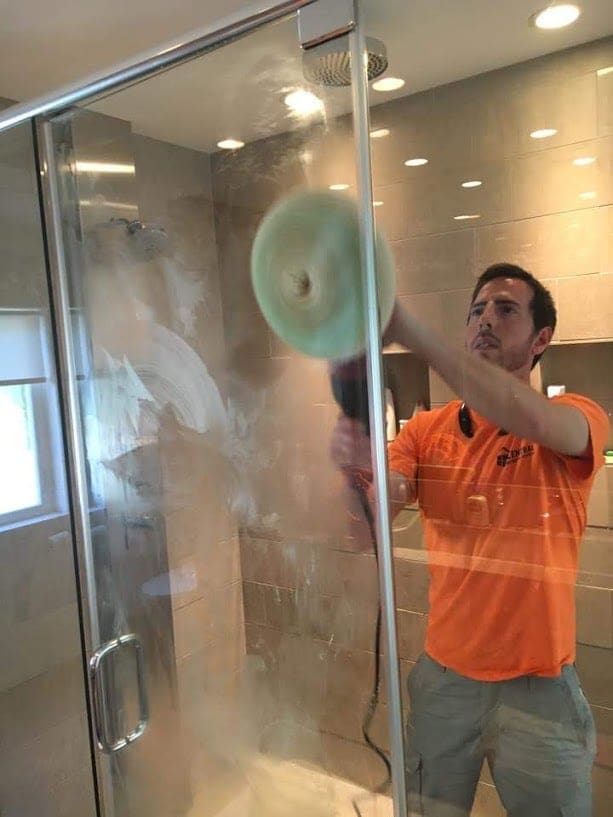 Technician in an orange shirt using a rotary buffing tool to restore or polish a glass shower door surface.