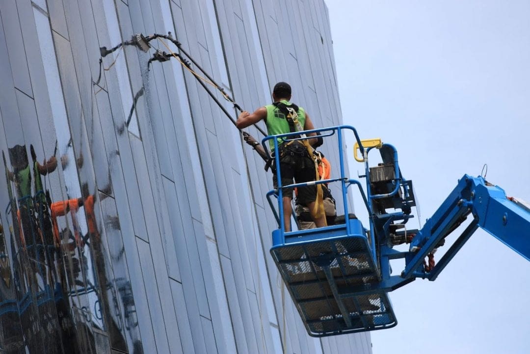 Technician on a blue cherry picker cleaning the reflective glass facade of a modern building under a clear sky.