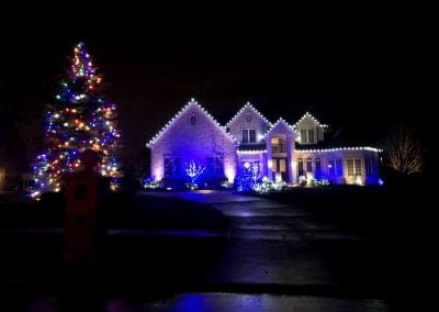 Large brick home at night decorated with cool white roofline lights and blue accent lighting on shrubs, with a tall outdoor Christmas tree glowing in multicolored lights on the left side of the yard.